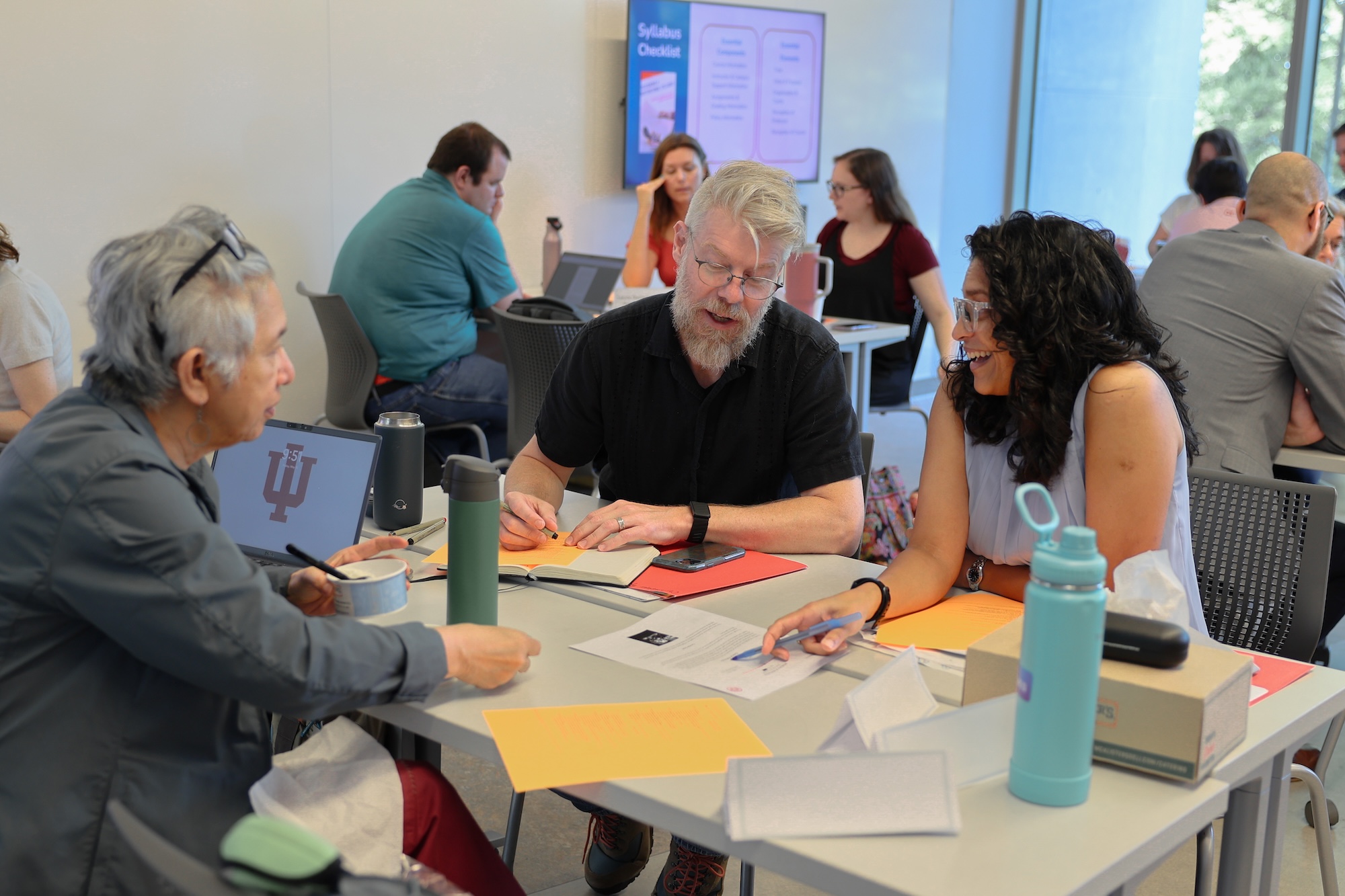 Three people sit at a table, discussing a worksheet. Other tables of participants are in the background